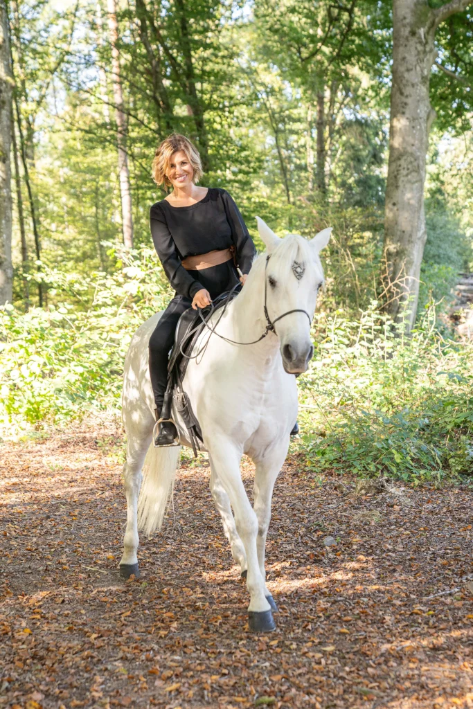 Frau auf einem weissen Pferd reitet durch den Wald
